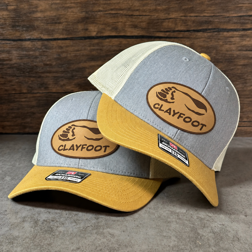 Digital artwork preview of the Clayfoot logo as a two-tone engraved oval patch on non-camouflage Richardson 115 hats; supplier proof pending